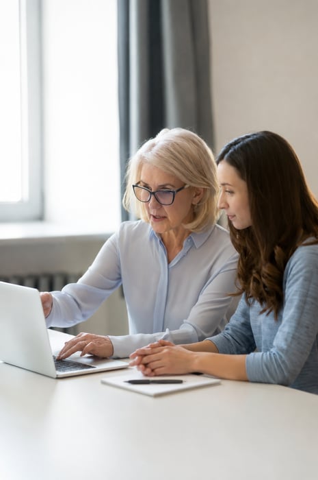 Two women looking at a laptop together at a table, one pointing at the screen as they review something side by side.