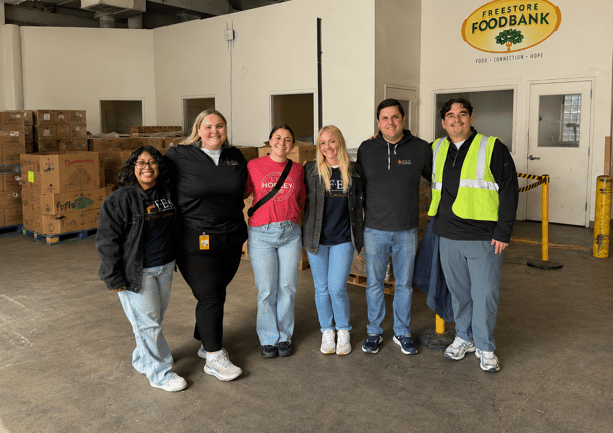 A group of FEG volunteers posing together in front of the Freestore Foodbank sign in a warehouse stocked with donated goods.