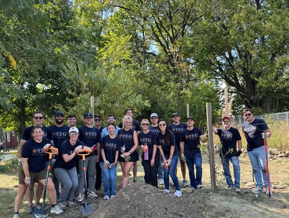 A large group of FEG employees in matching "FEG in the Community" t-shirts posing with shovels and gardening tools at an outdoor volunteer event.