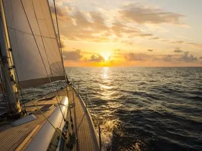 A sailboat with white sails gliding across open water toward a golden sunset on the horizon.