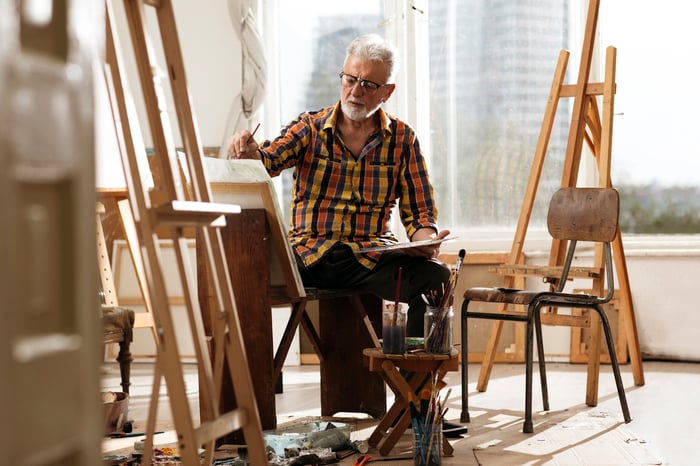 An older man in a plaid shirt painting at an easel in a sunlit studio surrounded by canvases, brushes, and easels.