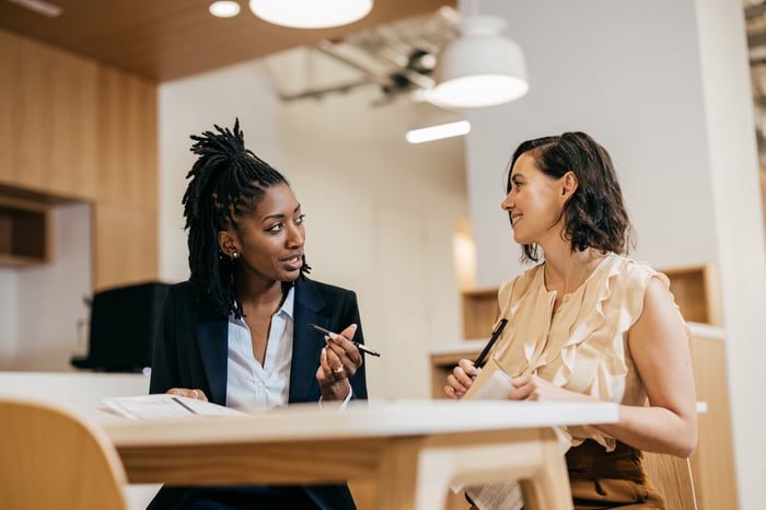 Two women in conversation at a desk in a bright office, one gesturing with a pen while the other listens and smiles.