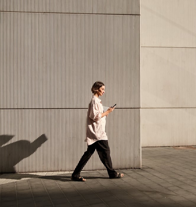 Woman in a light shirt and dark pants walking past a ribbed concrete wall, looking at her phone while holding a laptop in warm sunlight.