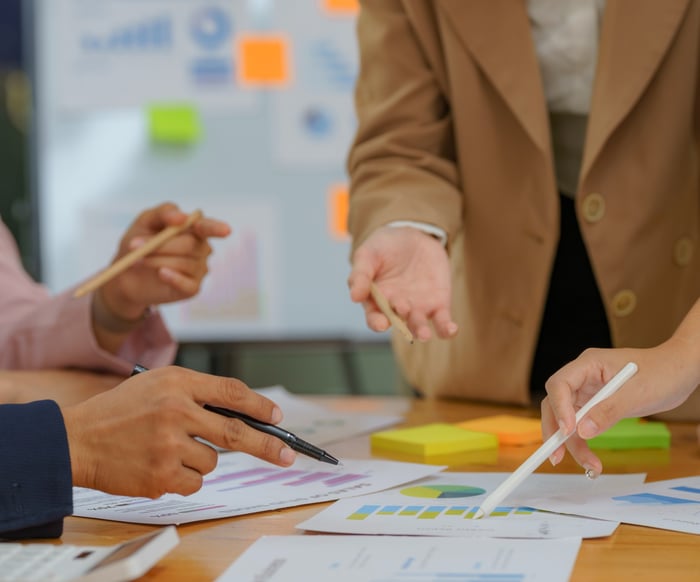 Colleagues collaborating around a table covered with colorful charts and sticky notes, gesturing as they discuss data during a business meeting.