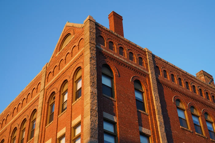 Corner of a historic red brick building with arched windows photographed from below against a clear blue sky in warm afternoon light.