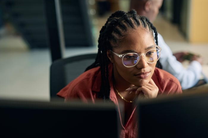 A woman with braids and clear-framed glasses studying a monitor intently, resting her chin on her hand in an office setting.