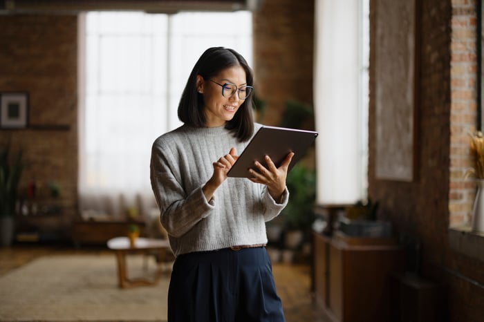 A woman in glasses and a gray sweater smiling while using a tablet in a bright, brick-walled loft space.
