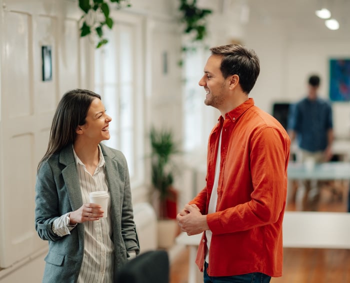 Two colleagues laughing and chatting casually in a bright office, one holding a coffee cup.Two colleagues laughing and chatting casually in a bright office, one holding a coffee cup.