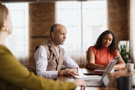 Colleagues in a meeting around a wooden table with a laptop and notes, a man in a plaid vest listening while a woman takes notes.