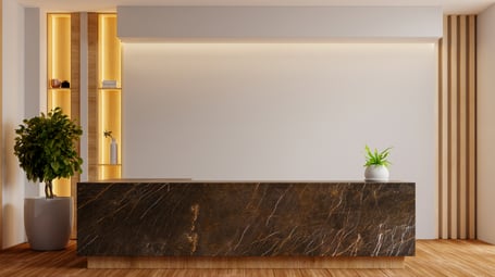 A modern reception area with a dark marble front desk, warm wood accents, backlit shelving, and potted plants in a minimalist lobby.