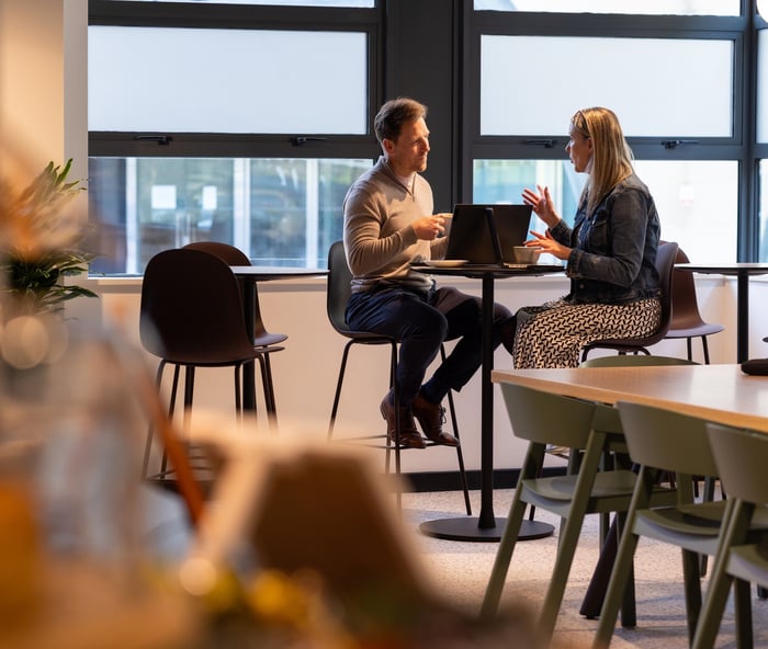 Two colleagues having an informal discussion over a laptop at a small table in a bright, modern office café.