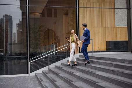 Two professionals talking on the steps of a modern glass office building in an urban setting, one holding a phone and the other carrying a laptop.Two professionals talking on the steps of a modern glass office building in an urban setting, one holding a phone and the other carrying a laptop.