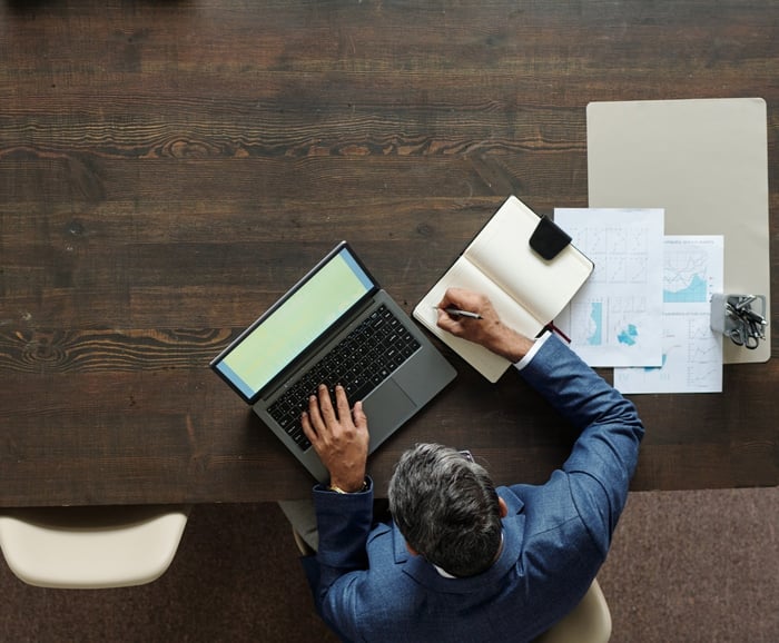 Overhead view of a man in a blue suit working at a wooden desk, typing on a laptop while taking notes beside printed charts and graphs.