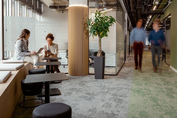 A modern open office lounge with two women working on laptops in a seating nook and two colleagues walking past in the background.