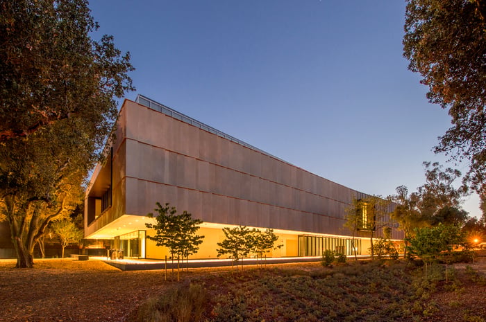 Modern museum or institutional building with a paneled facade and warmly lit glass ground floor, photographed at dusk among mature oak trees.
