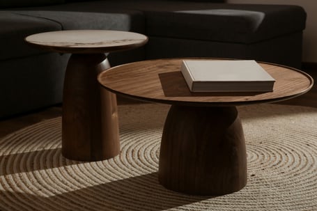 Two round pedestal coffee tables in walnut and marble on a woven rug, with a book resting on one, in a warmly lit living room.