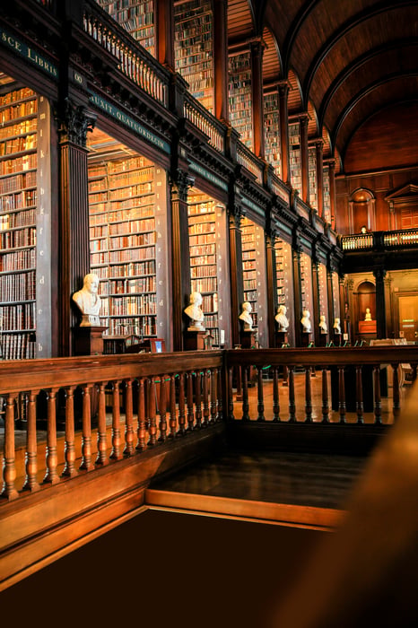 Interior of a grand historic library with floor-to-ceiling dark wood bookshelves, marble busts, and a barrel-vaulted ceiling.