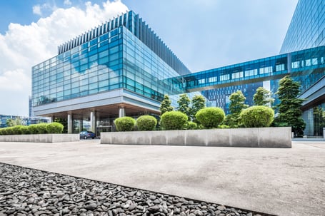 A modern blue-glass office building with a connecting skybridge, fronted by manicured shrubs and an empty plaza under a bright sky.