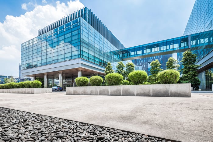 A modern blue-glass office building with a connecting skybridge, fronted by manicured shrubs and an empty plaza under a bright sky.