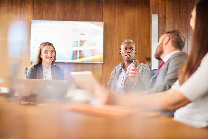 Colleagues in a wood-paneled boardroom during a meeting, with a presentation screen in the background and a woman speaking to the group.