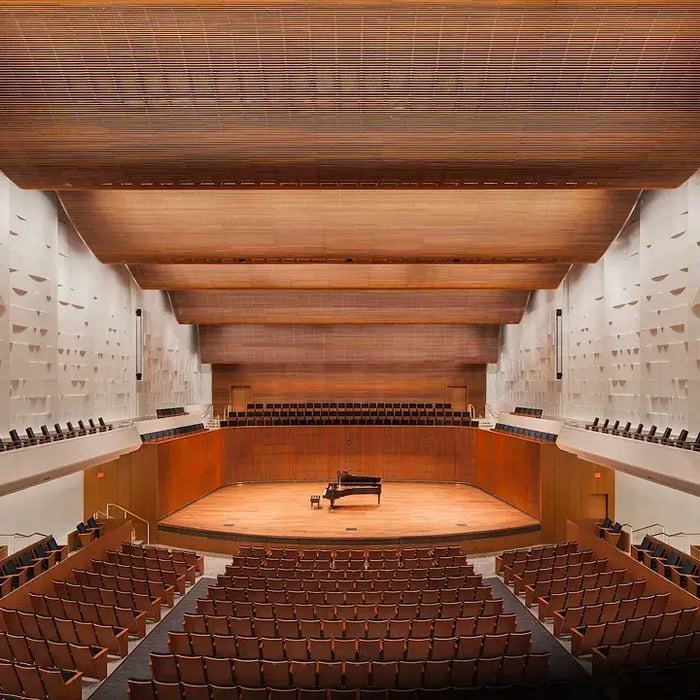 Empty concert hall with warm wood paneling and tiered seating, a grand piano alone on the stage.